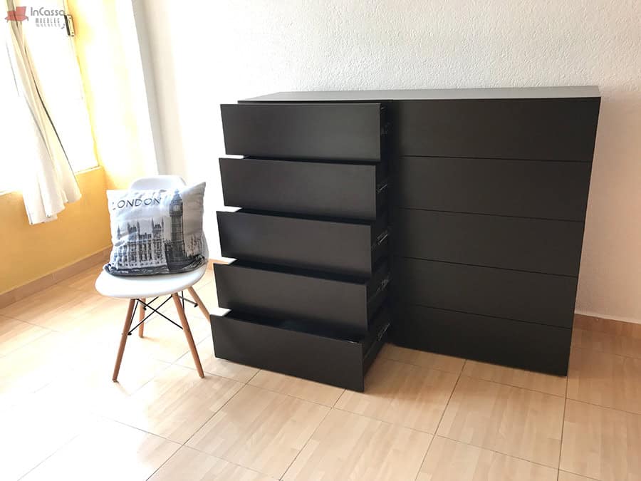 La imagen muestra una toma frontal de la cajonera Tel Aviv en acabado chocolate mate, con todos los cajones del lado izquierdo completamente abiertos. A su lado, una silla decorativa con cojines y una ventana que aporta luz natural, resaltan la funcionalidad y estilo del mueble.