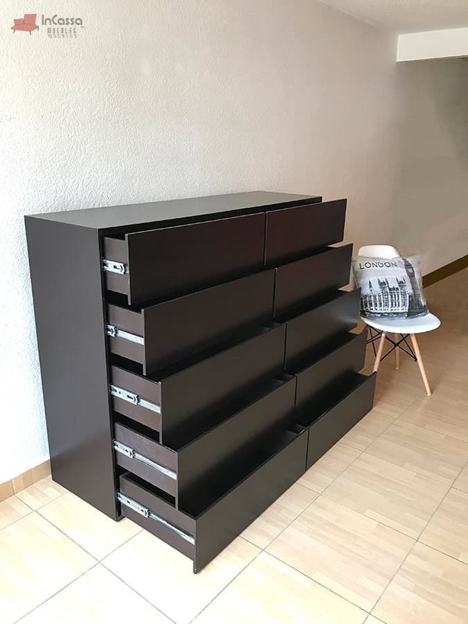 Una fotografía tomada en ángulo lateral desde el pasillo muestra la cajonera Tel Aviv en color chocolate mate con todos sus cajones abiertos. Se aprecian los rieles metálicos de extensión y una silla decorativa a un costado, en un ambiente minimalista de interior.