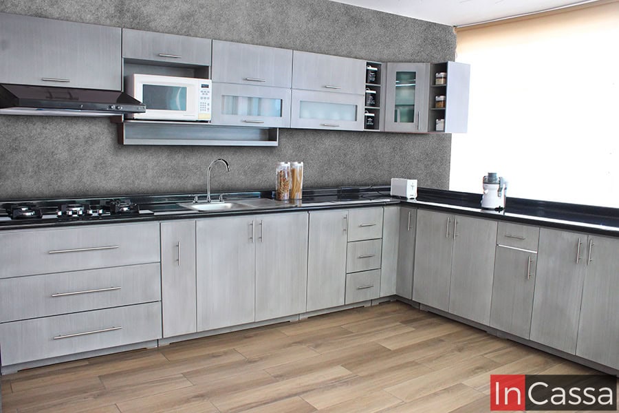 En esta imagen se aprecia la Cocina Tailandia en escuadra, con acabado en gris cenizo claro. Fabricada en MDF, integra cubierta de formaica estilo mármol negro, múltiples gabinetes y cajones, ideal para parrilla y optimizar cada espacio.