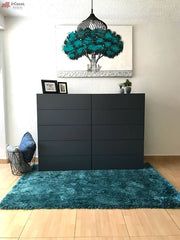 La imagen muestra una cajonera en color gris oxford con 8 cajones amplios, presentada en un entorno decorado con alfombra verde esmeralda, cuadros y luz natural. Su diseño minimalista resalta en espacios modernos y bien iluminados.