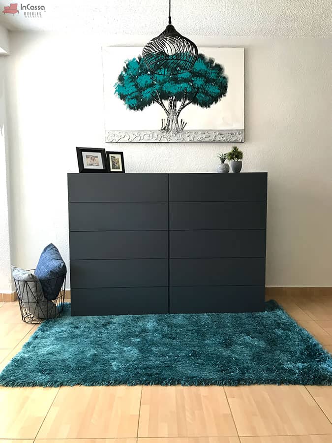 La imagen muestra una cajonera en color gris oxford con 8 cajones amplios, presentada en un entorno decorado con alfombra verde esmeralda, cuadros y luz natural. Su diseño minimalista resalta en espacios modernos y bien iluminados.