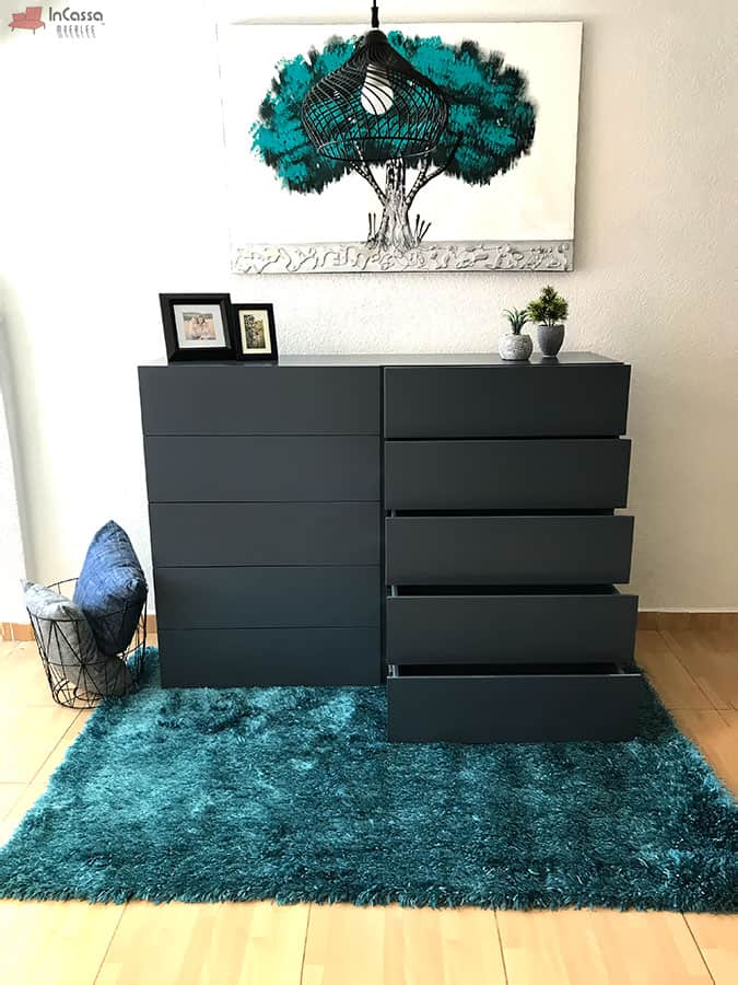 La fotografía destaca la funcionalidad de la cajonera Tel Aviv en tono gris oxford, mostrando cinco de sus cajones abiertos. El mueble se encuentra ambientado con una alfombra verde esmeralda, decoración mural y luz natural, ideal para dormitorios modernos.