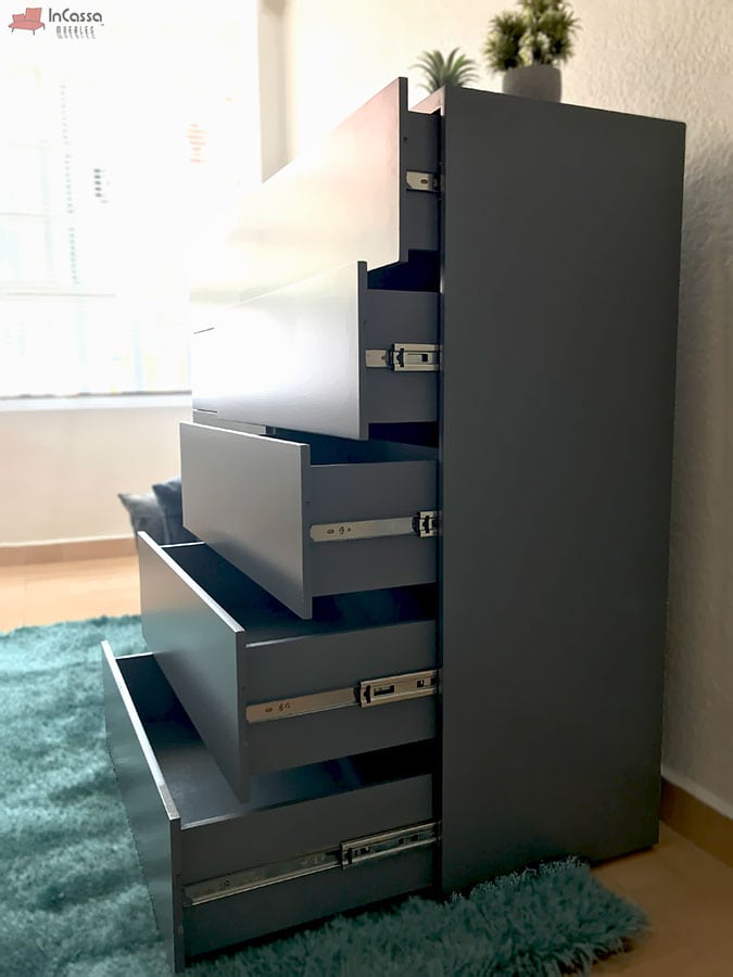 Una toma diagonal revela el lateral izquierdo de la cajonera Tel Aviv en tono gris oxford, destacando la apertura de sus cajones con correderas metálicas visibles. Se aprecia en una habitación luminosa con alfombra verde y detalles decorativos modernos.