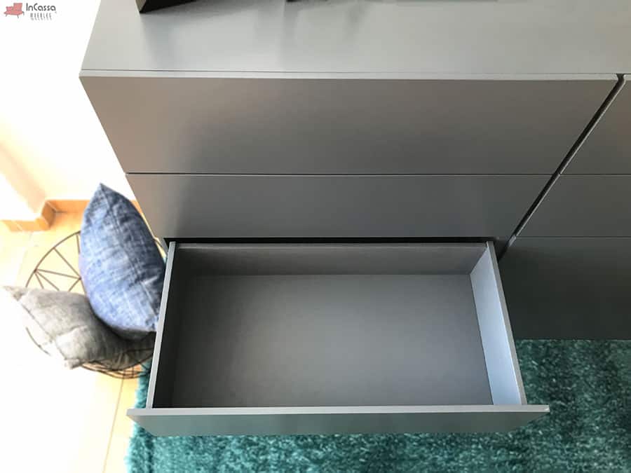 La fotografía muestra desde arriba uno de los cajones abiertos de la cajonera Tel Aviv, revelando su interior espacioso en acabado gris oxford. El entorno incluye una alfombra turquesa y cojines decorativos, aportando calidez y estilo al ambiente.