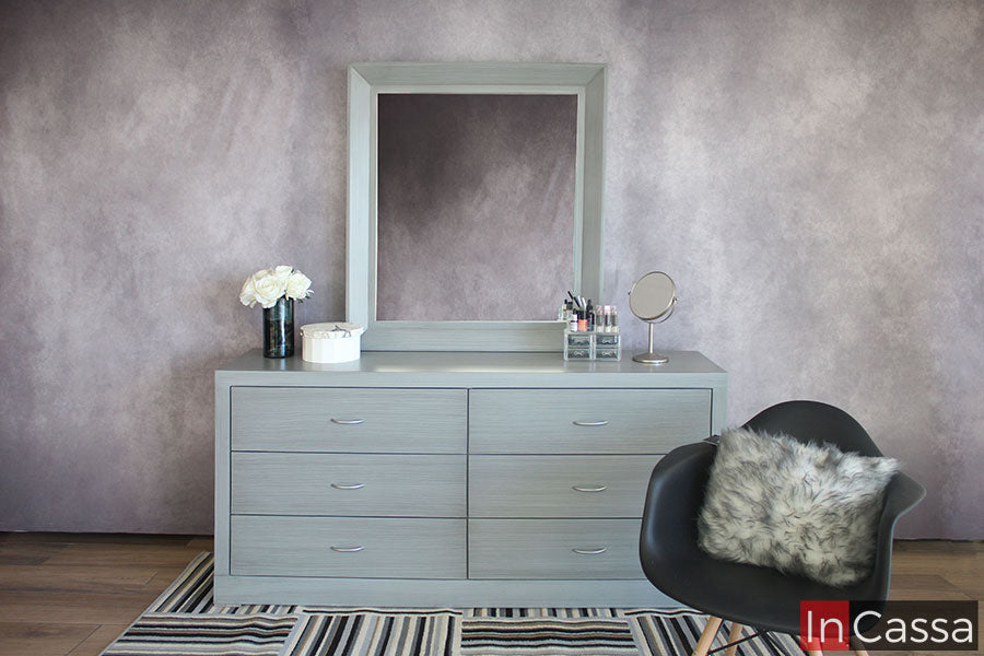 Tocador contemporáneo en acabado gris cenizo claro, el mueble consta de un tocador de 6 cajones y un espejo rectangular, todo instalado en una pared gris.