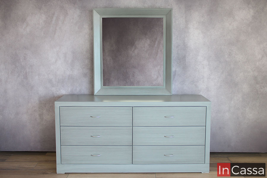 Imagen completa del tocador de estilo contemporáneo, el cuál incluye seis cajones iguales en su amplio diseño así como un espejo rectangular. Todas y cada una de las partes que conforman el mueble están pintadas en un tono cenizo claro de acabado laqueado.