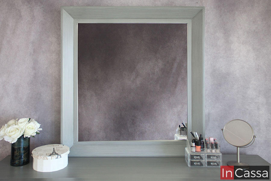 La foto se concentra completamente en el espejo del tocador, cuyo marco mantiene el acabado cenizo claro que se puede encontrar en la totalidad del mueble. Además, también se puede apreciar la cubierta del tocador, la cuál es decorada por varios utensilios y maquillajes a fin de ejemplificar su longitud.