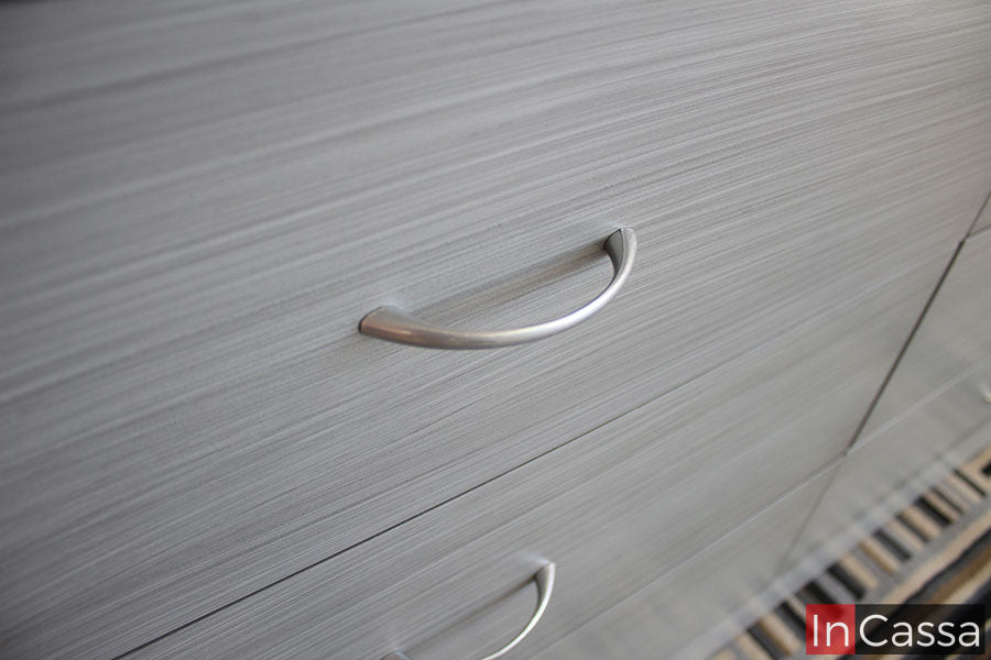 La imagen toma un enfoque cercano a las jaladeras integradas en los cajones, las cuales podemos ver son completamente metálicas con forma de arco. Adicionalmente se puede apreciar de manera más cercana el acabado cenizo claro laqueado en algunos de los cajones.