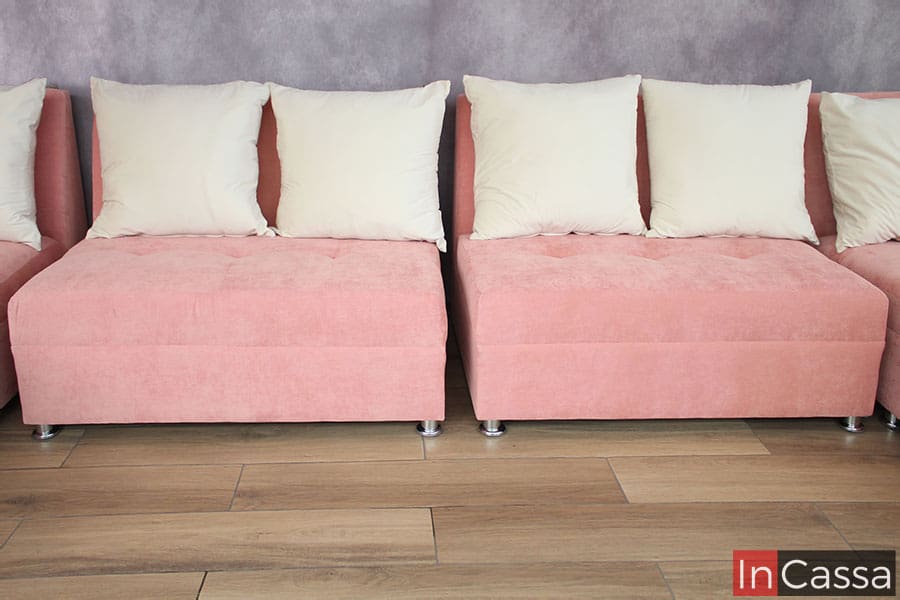 La foto se enfoca en mostrar de frente los dos love seat, tapizados en suede rosa, que conforman parte de la sala. Ambos love seat están separados por un pequeño espacio con el fin de ejemplificar sus dimensiones de manera individual, lo cual se resalta más fácilmente gracias a los cojines hueso que los acompañan.