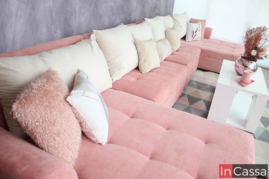 Vemos una toma lateral más completa de la sala suede rosa, visualizando más de cerca el gran asiento del chaise long izquierdo así como el del resto de los módulos de manera general, resaltando los seis cojines grandes en suede hueso y los cojines pequeños decorativos en colores rosa, blanco y hueso (cojines decorativos no incluidos).