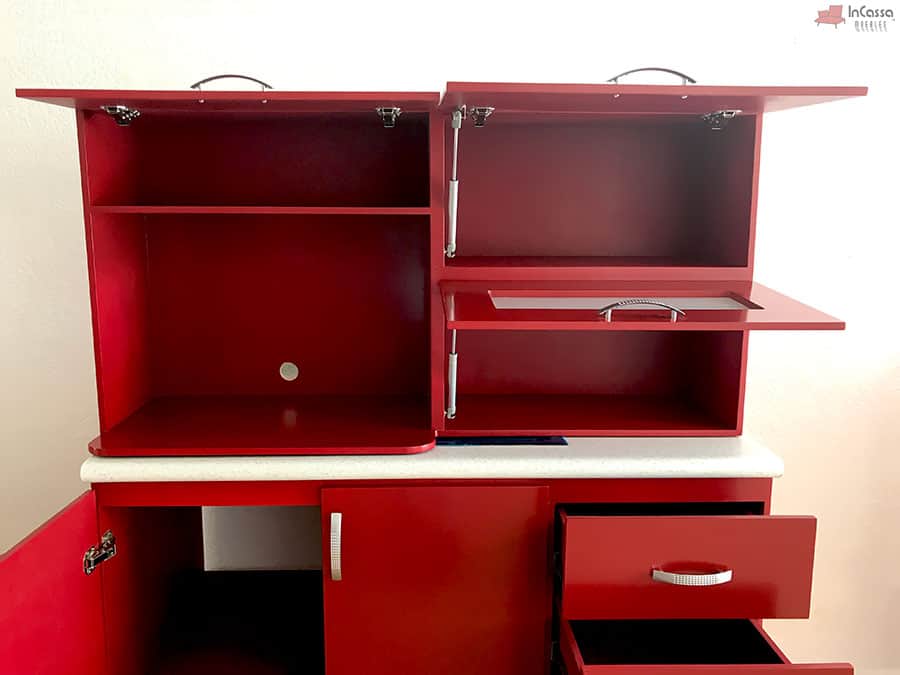 Una toma de acercamiento al módulo superior muestra alacenas con apertura vertical asistida por pistones neumáticos y compartimentos amplios para almacenar utensilios o despensa. También se observan cajones parcialmente abiertos y el espacio interior del módulo bajo tarja. El acabado rojo quemado aporta un estilo moderno y llamativo.