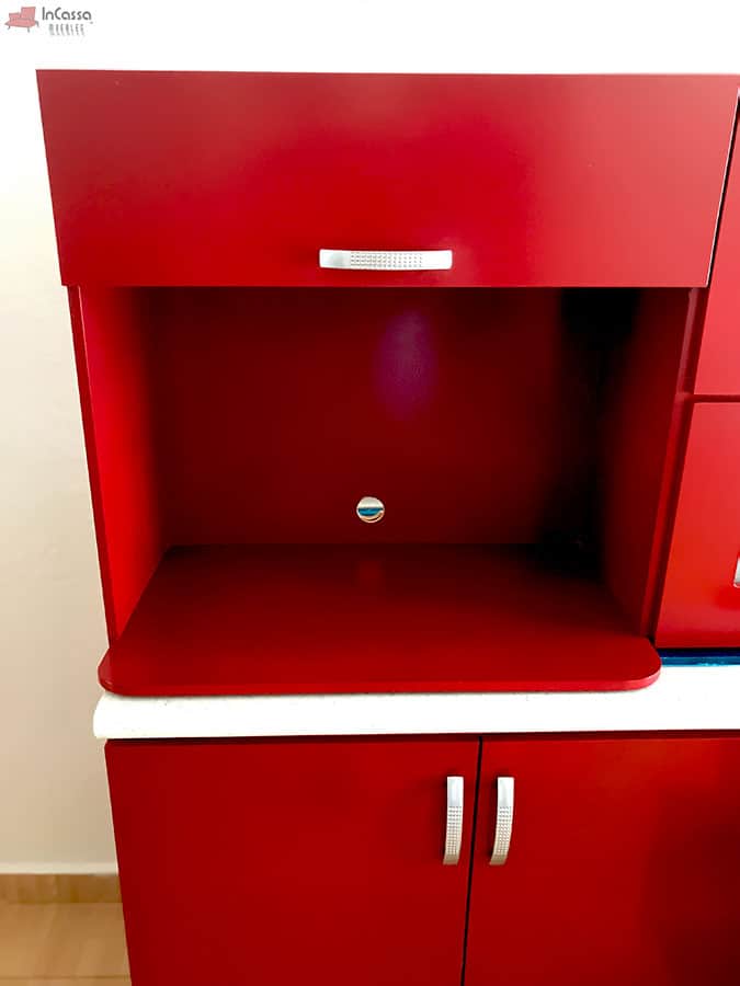 La imagen muestra un compartimento específico para microondas, equipado con orificio trasero para cables. Destaca el acabado rojo quemado del mueble, acompañado por jaladeras metálicas modernas.