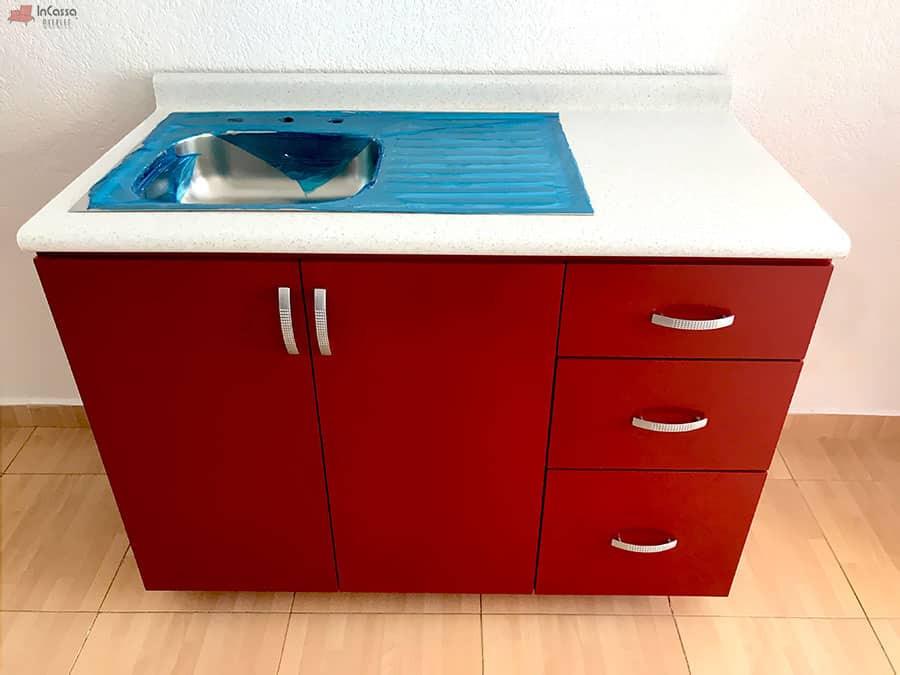 En esta imagen se aprecia el módulo de tarja en color rojo quemado, con cubierta de formaica blanco fresh y tarja de acero inoxidable. El diseño incluye tres cajones funcionales y un compartimento doble con jaladeras modernas en acabado metálico.