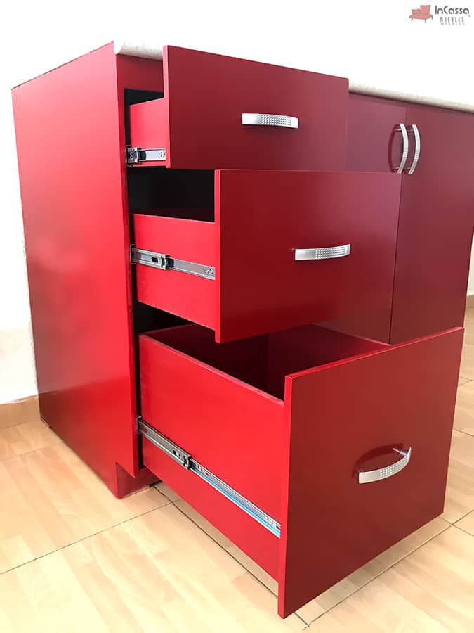 La imagen muestra un acercamiento de los tres cajones del módulo inferior, completamente abiertos. Se observa el sistema de correderas metálicas de extensión y las elegantes jaladeras texturizadas en acabado metálico. El color rojo quemado resalta el estilo moderno del diseño.