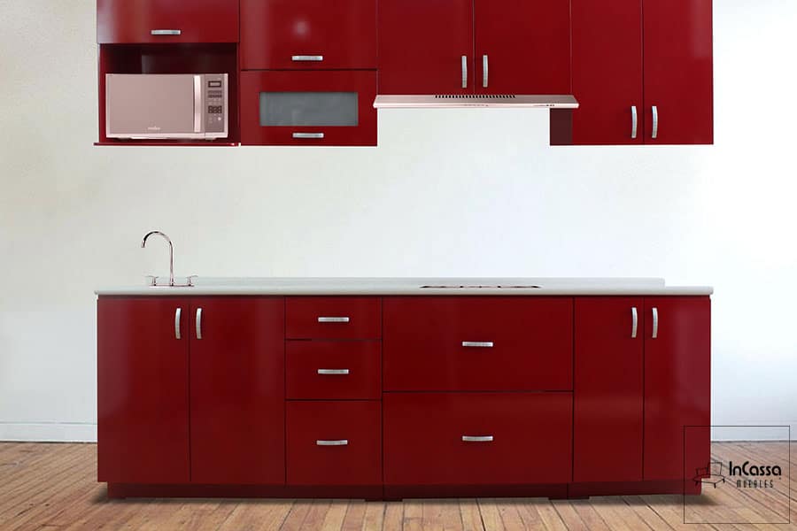 Una toma frontal de la cocina, en la que destaca su gran numero de compartimentos incluyendo un espacio porta microondas incluido en el módulo sobre tarja. La cocina está pintada en su totalidad en color borgoña brillante, mientras que su cubierta de formica tiene un acabado estilo sal y pimienta.