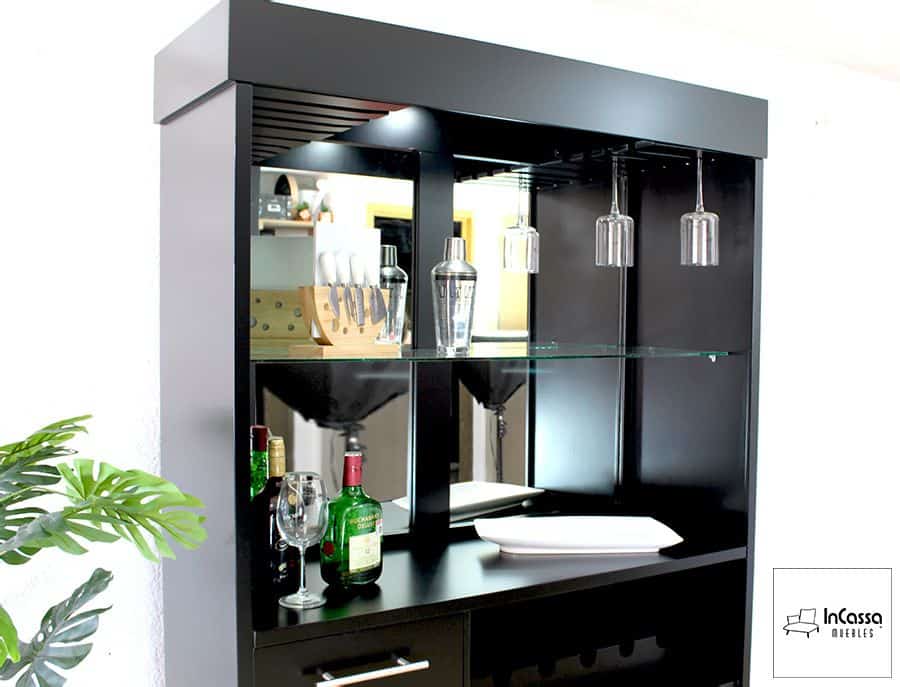 La imagen resalta el acabado chocolate del mueble y su espejo posterior que amplía visualmente el espacio. Desde esta perspectiva se aprecian el estante de cristal flotante, copas suspendidas en riel metálico y la iluminación LED superior, ideal para crear una atmósfera elegante.