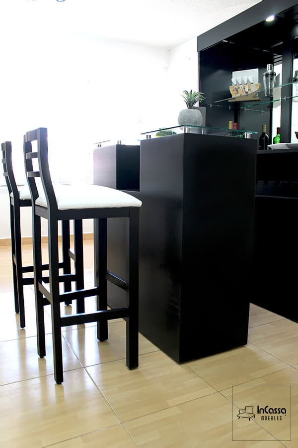 Una toma en ángulo bajo que resalta el diseño estilizado de la barra modelo TUCSON en color chocolate mate, con cubierta de cristal templado sostenida por soportes metálicos circulares. Al frente se aprecian dos bancos altos con estructura del mismo tono y asiento tapizado en color blanco, creando un contraste elegante y moderno. El fondo muestra la contra barra con estantes, cajones, copero y repisa de cristal.