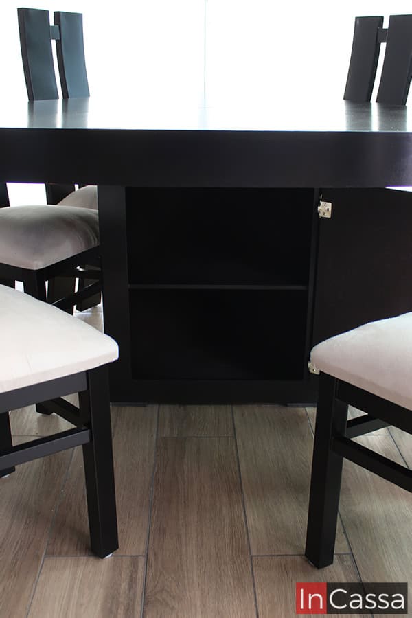 Toma frontal que muestra el gabinete central en el pedestal de la mesa en color chocolate, con la puerta abierta revelando un entrepaño interior para almacenamiento. Las sillas con cojines claros enmarcan el mueble, destacando sobre el piso tipo madera.