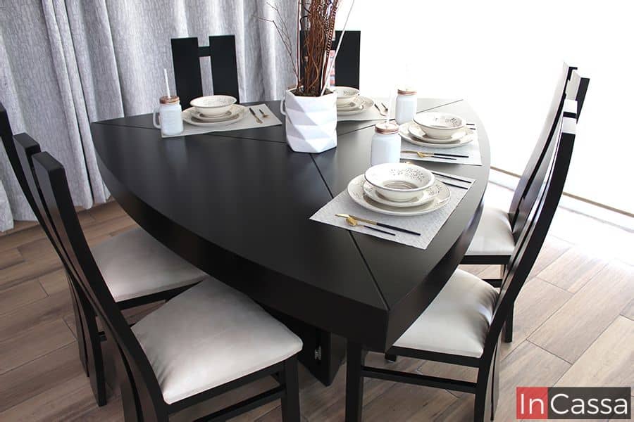 Imagen tomada en ángulo superior lateral que muestra el diseño triangular de la mesa chocolate, rodeada por seis sillas tapizadas en suede hueso. La disposición de la vajilla y el florero central aportan elegancia y simetría al conjunto.