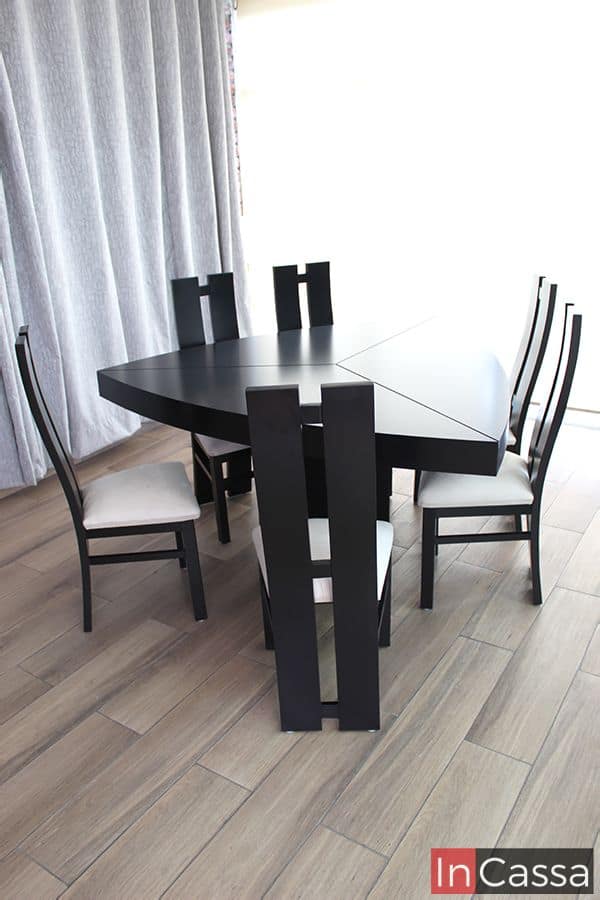 Fotografía lateral que resalta la estructura geométrica de la mesa chocolate en forma triangular, acompañada por seis sillas de respaldo alto y tapizado suede hueso. El contraste entre el acabado oscuro del mueble y el piso tipo madera realza su diseño contemporáneo.