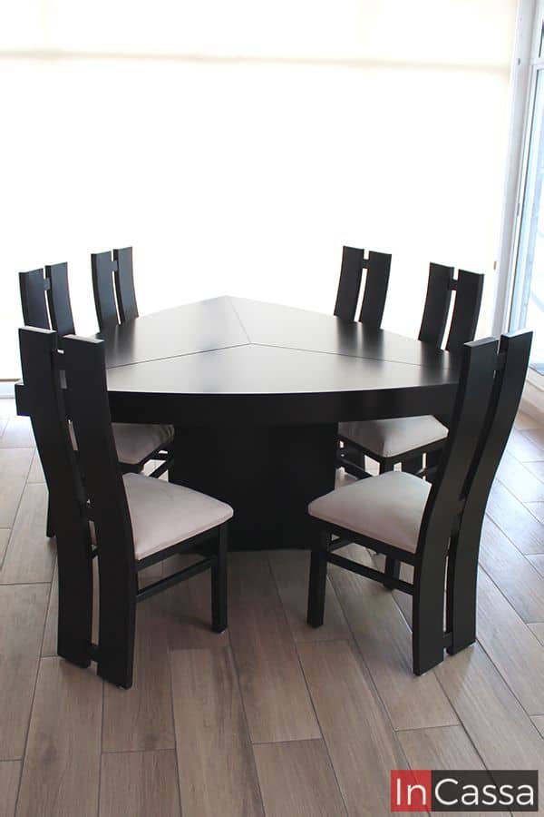 Imagen frontal que muestra la elegante mesa chocolate de forma triangular, acompañada por seis sillas de respaldo alto y cojines tapizados en suede hueso. El contraste con el piso tipo madera y la luz natural resalta su diseño moderno y sofisticado.