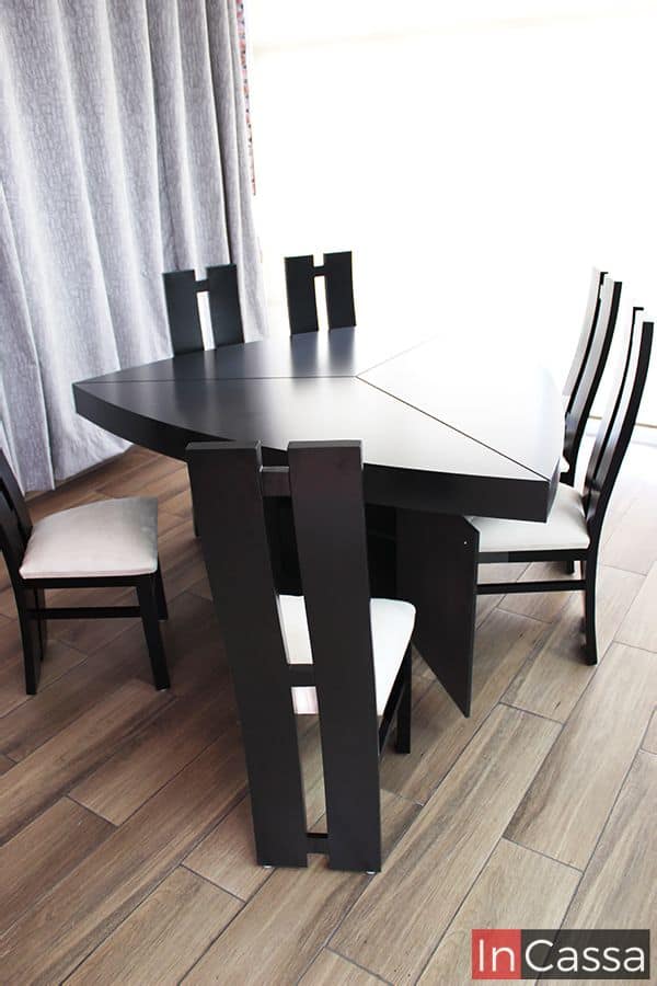 Toma lateral que resalta el diseño triangular de la mesa en color chocolate, mostrando una de sus puertas abiertas para revelar el espacio de almacenamiento interior. Las sillas de respaldo alto con cojines de suede hueso contrastan con el piso tipo madera y la cortina gris de fondo.