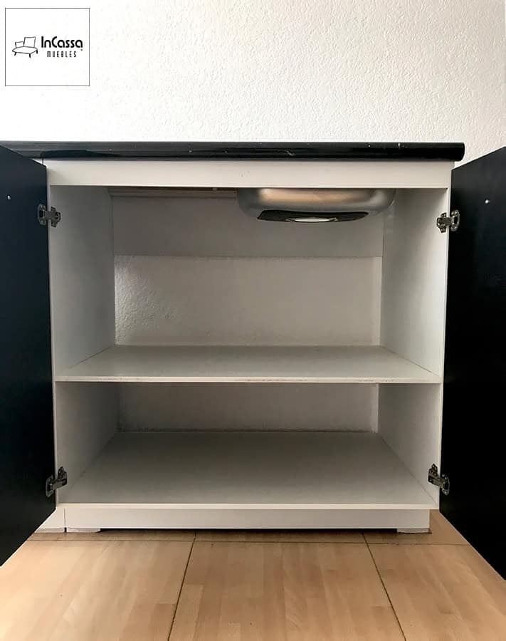 Una toma frontal que muestra el interior del módulo bajo ubicado justo debajo de la tarja, con estructura blanca y doble entrepaño amplio para organización. Se aprecia la base metálica de la tarja montada en la parte superior, junto a las puertas en tono chocolate abiertas, destacando su capacidad y fácil acceso.