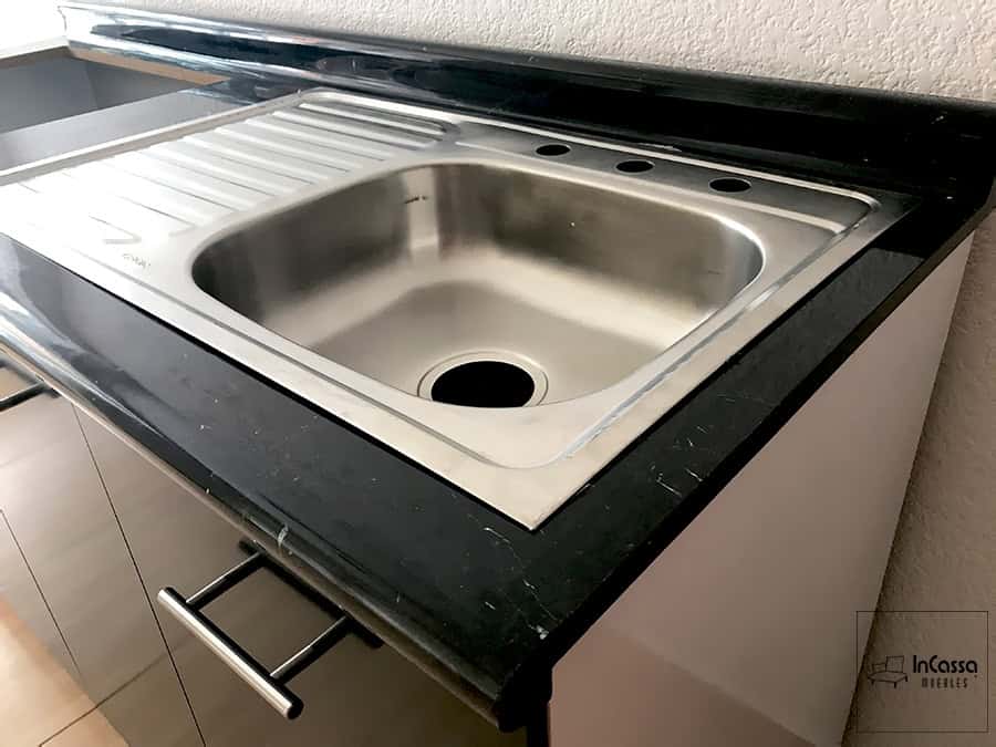 Una toma en ángulo que resalta la tarja de acero inoxidable perfectamente instalada sobre la cubierta de formaica estilo mármol negro. Se observan las orillas redondeadas y el escurridor integrado, junto con los frentes chocolate y jaladeras metálicas que aportan un acabado moderno y elegante.