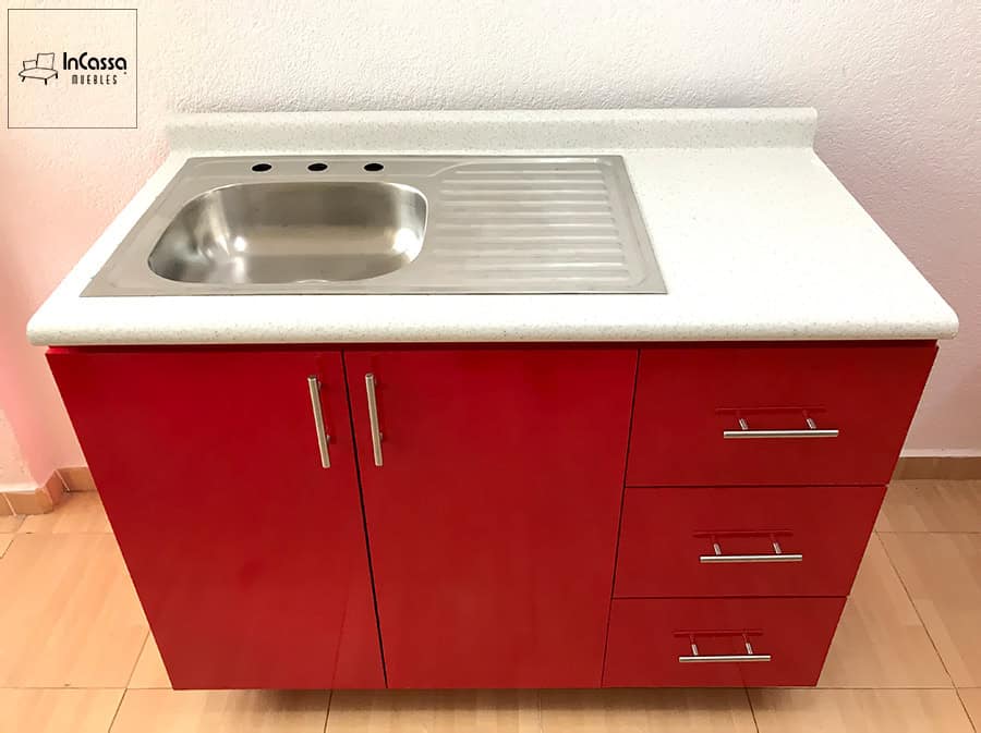 Una fotografía del módulo inferior de la Cocina Finlandia en rojo vivo, con tarja de acero inoxidable y escurridor integrado sobre la cubierta de formaica blanco fresh. Destacan los cajones metálicos y puertas con tiradores modernos.