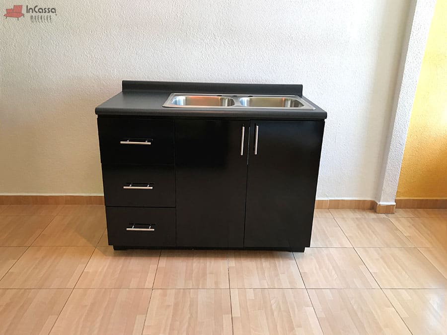 La foto presenta el módulo inferior de la Cocina Integral Irlanda para Estufa en color negro brillante, equipado con tarja doble de acero inoxidable y cubierta de formaica estilo mármol negro. A la izquierda se aprecian tres cajoneras con correderas metálicas, mientras que al centro destacan puertas con tiradores modernos que ofrecen amplio espacio de guardado.