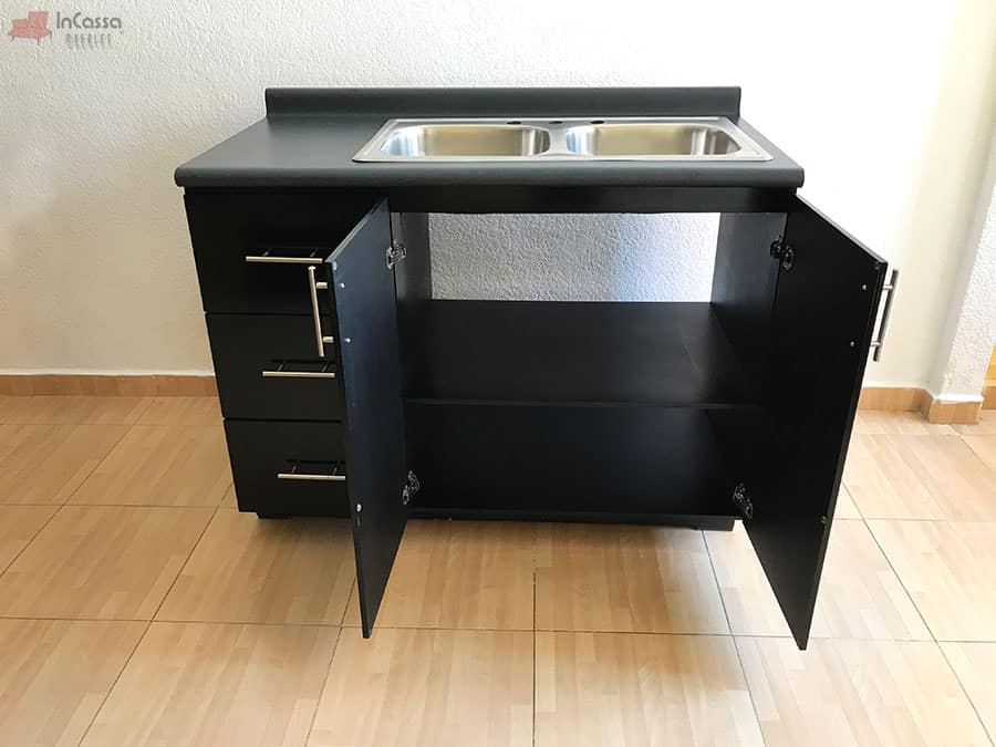 La foto muestra el módulo inferior de la Cocina Integral Irlanda para Estufa en color negro brillante, con tarja doble de acero inoxidable y puertas abiertas que revelan un amplio espacio interno dividido por repisa. A la izquierda, se aprecian tres cajoneras con herrajes metálicos modernos, ideales para mantener utensilios organizados.