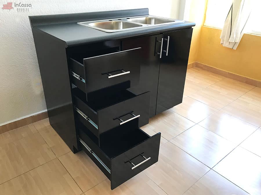 Una toma lateral del módulo inferior de la Cocina Integral Irlanda para Estufa en negro brillante, con tarja doble de acero inoxidable sobre cubierta en mármol negro. Se aprecian los tres cajones abiertos que destacan su sistema corredizo metálico de extensión completa, diseñadas para ofrecer resistencia, funcionalidad y un estilo moderno en la organización.