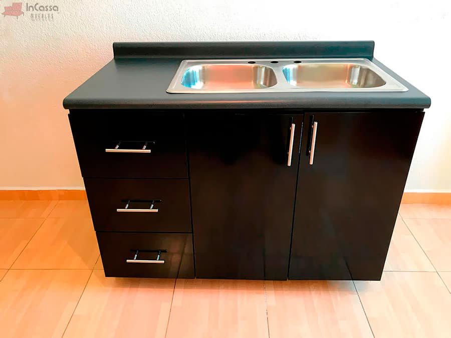 Se observa el módulo inferior de la Cocina Integral Irlanda para Estufa en acabado negro brillante, con tarja doble de acero inoxidable y cubierta de formaica estilo mármol negro. A la izquierda sobresalen tres cajones amplios con jaladeras metálicas modernas, mientras que al centro las puertas ofrecen un espacio funcional para organización y almacenamiento.