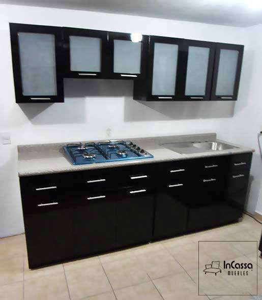 La imagen muestra una cocina integral de estilo contemporáneo, con gabinetes en tono negro brillante y puertas superiores con vidrio esmerilado. Con una parrilla empotrada y una tarja de acero inoxidable sobre una cubierta de formaica en estilo mármol escalona. Las jaladeras metálicas alargadas realzan su diseño elegante y funcional.