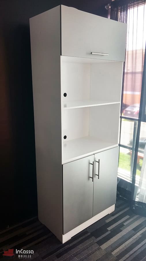 Se presenta el mueble para horno en acabado blanco y silver visto en ángulo lateral, con puertas superiores e inferiores cerradas y estantes centrales vacíos. Las perforaciones traseras para cables y las jaladeras metálicas rectas complementan su diseño funcional y contemporáneo.