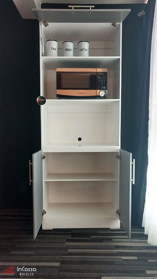 La foto presenta el mueble para horno Windsor en tonos blanco y silver visto de frente, con la puerta superior levantada y las inferiores abiertas, revelando amplios compartimentos internos. En el estante intermedio destaca un horno de microondas y en el superior tres recipientes metálicos rotulados para té, azúcar y café, con iluminación natural proveniente del lateral derecho.