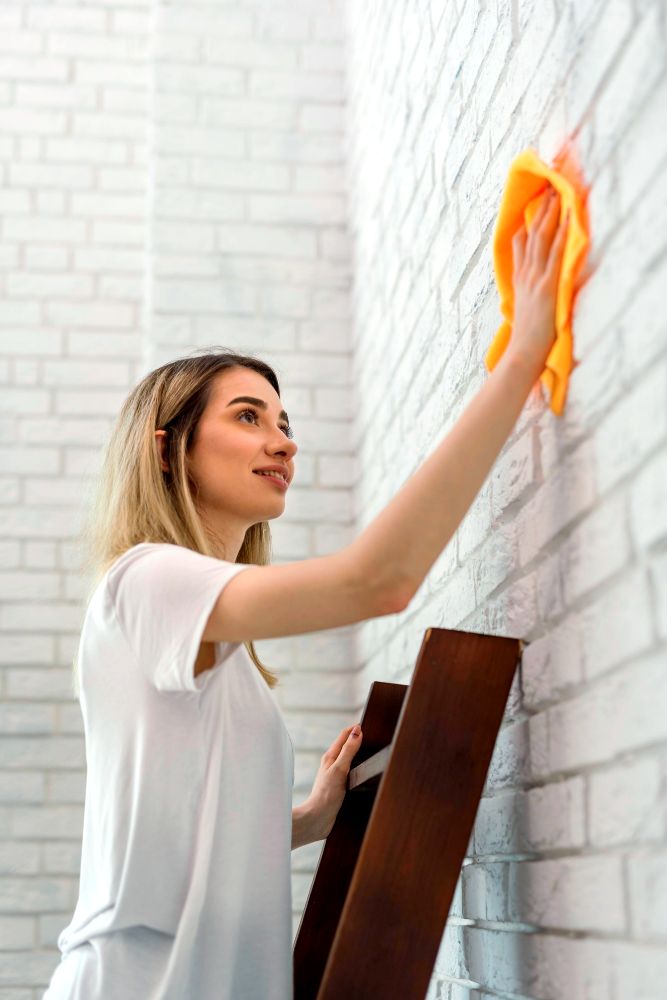 La imagen nos muestra a una mujer secando una pared con un paño seco, utilizando una escalera para acceder a la parte más alta y secando enteramente la pared.