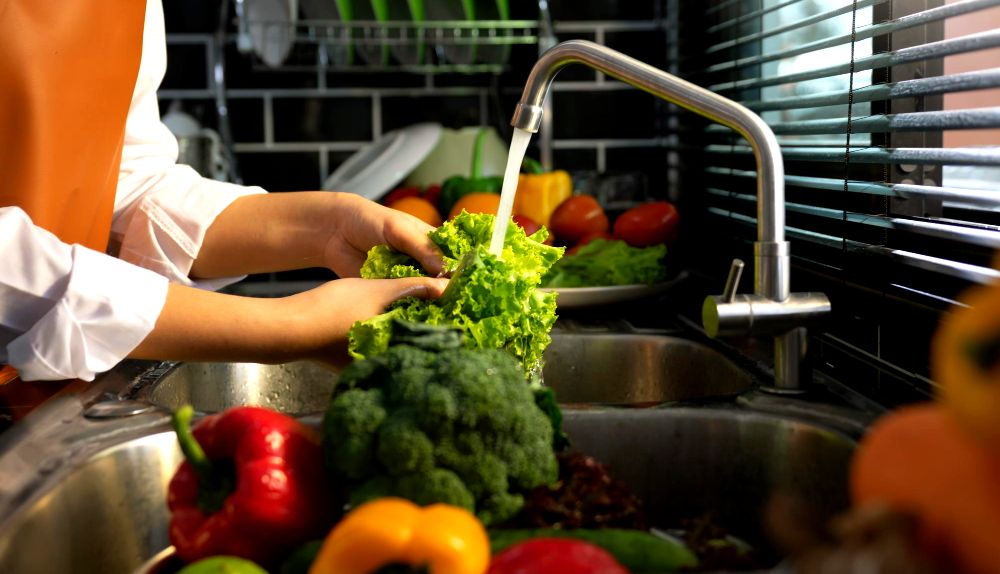 Persona lavando hojas de lechuga y otros vegetales bajo el grifo de la cocina, con un fondo de utensilios y alimentos.