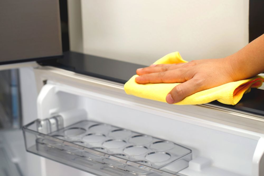 La imagen muestra a una persona limpiando cuidadosamente el interior de un refrigerador con un paño amarillo, asegurando la higiene del aparato.
