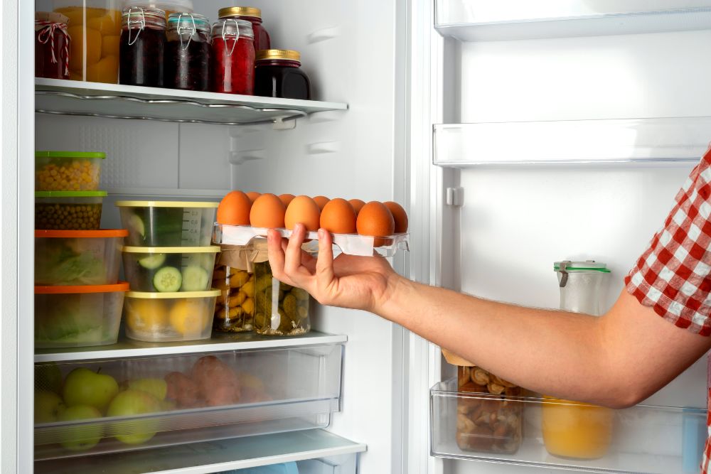 Persona colocando una bandeja de huevos en el estante de la puerta del refrigerador, con otros alimentos almacenados alrededor.