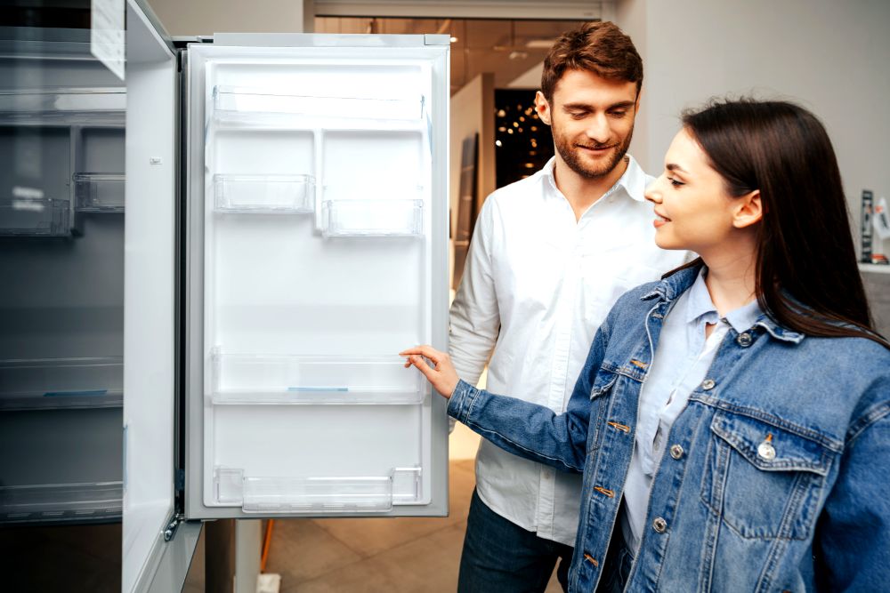 Pareja observando y evaluando un refrigerador vacío en una tienda de electrodomésticos, posiblemente considerando una compra.