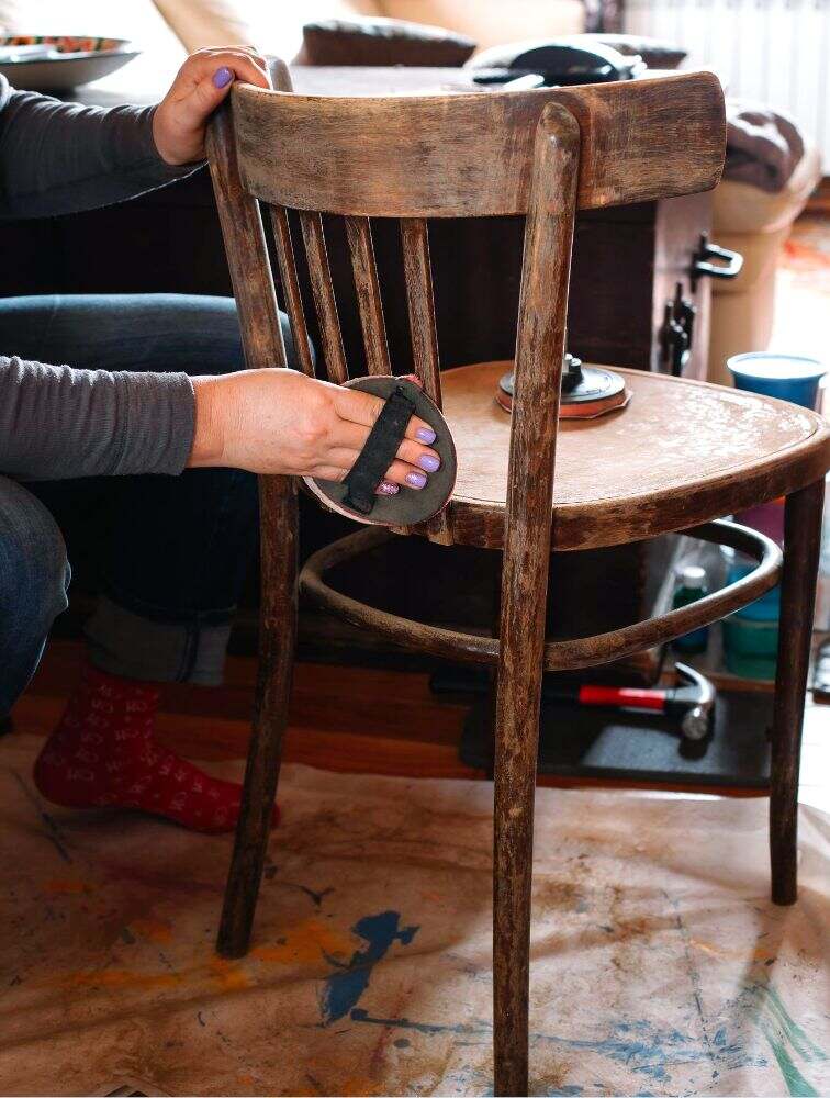 La foto muestra a una persona restaurando una pequeña silla de madera rústica, la cuál puede funcionar como un excelente articulo decorativo para espacios pequeños.