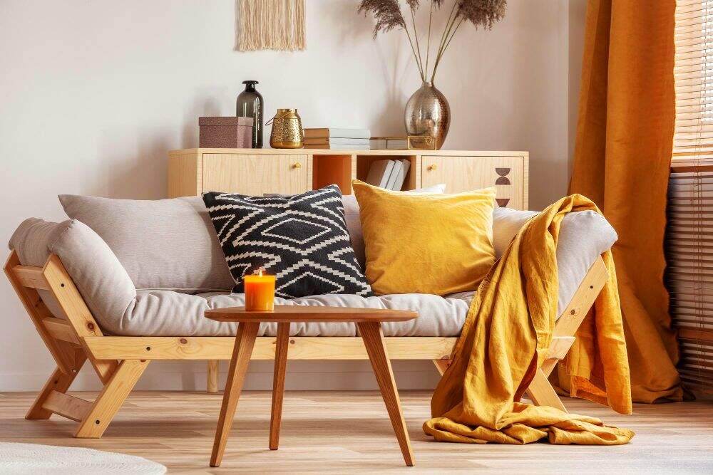 La foto presenta una estancia con una gran variedad de muebles minimalistas elaborados con madera natural y decorados por tejidos, plantas y velas de tonos cálidos dando como resultado un ambiente muy acogedor.