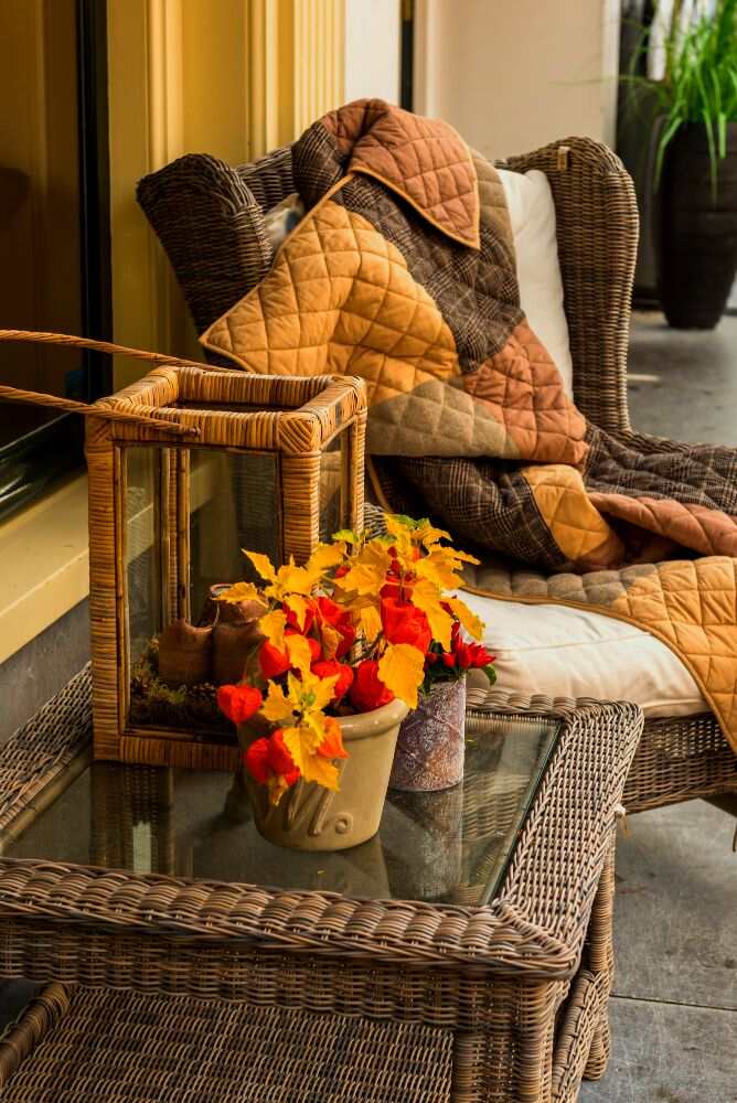 Un juego de sillón y mesa elaborados de ratan y adornados con articulos de diferentes tonos de naranjas entre los que se encuentran unas plantas decorativas, una manta y una lampara rústica.