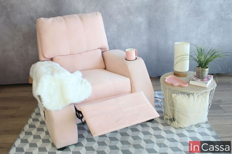 Una imagen del sillón reposet tapizado en suede rosa palo exhibido en una estancia con pared gris y piso de madera. El sillón se presenta con su reposapies extendido y con una taza en el porta vasos integrado al reposabrazos.