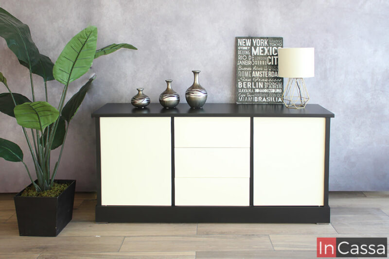 Bufetero de estilo minimalista en color negro con puertas y cajones blancos. El mueble se encuentra exhibido en una estancia con pared gris y piso de madera acompañado de una gran planta decorativa.