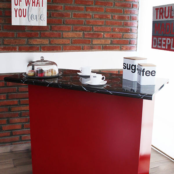 Barra minimalista de acabado rojo brillante con cubierta de formica estilo mármol negro, el mueble es exhibido en una estancia con pared de ladrillo, ventana completa y piso de madera.