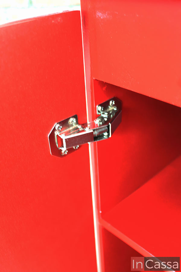 Se nos enseña un enfoque completo a una de las bisagras metálicas que conforman el sistema de apertura y cierre de cada puerta. También se nos ofrece una mejor vista de parte de los interiores, mismos que mantienen el mismo color rojo que la estructura externa del mueble.