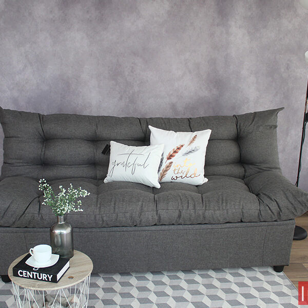 Sofá cama tapizado en tela lino mouse en acabado capitoneado completo, instalado frente a una pared gris en una habitación con piso de madera y adornado con 2 cojines blancos.