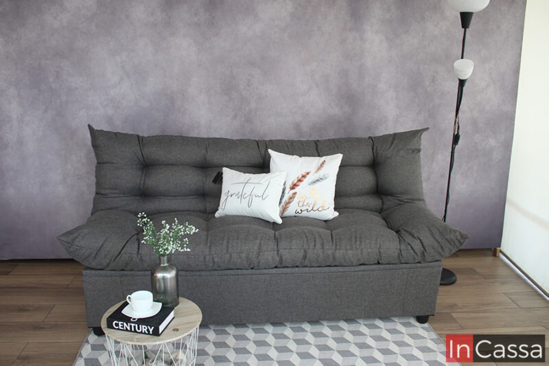 Sofá cama tapizado en tela lino mouse en acabado capitoneado completo, instalado frente a una pared gris en una habitación con piso de madera y adornado con 2 cojines blancos.